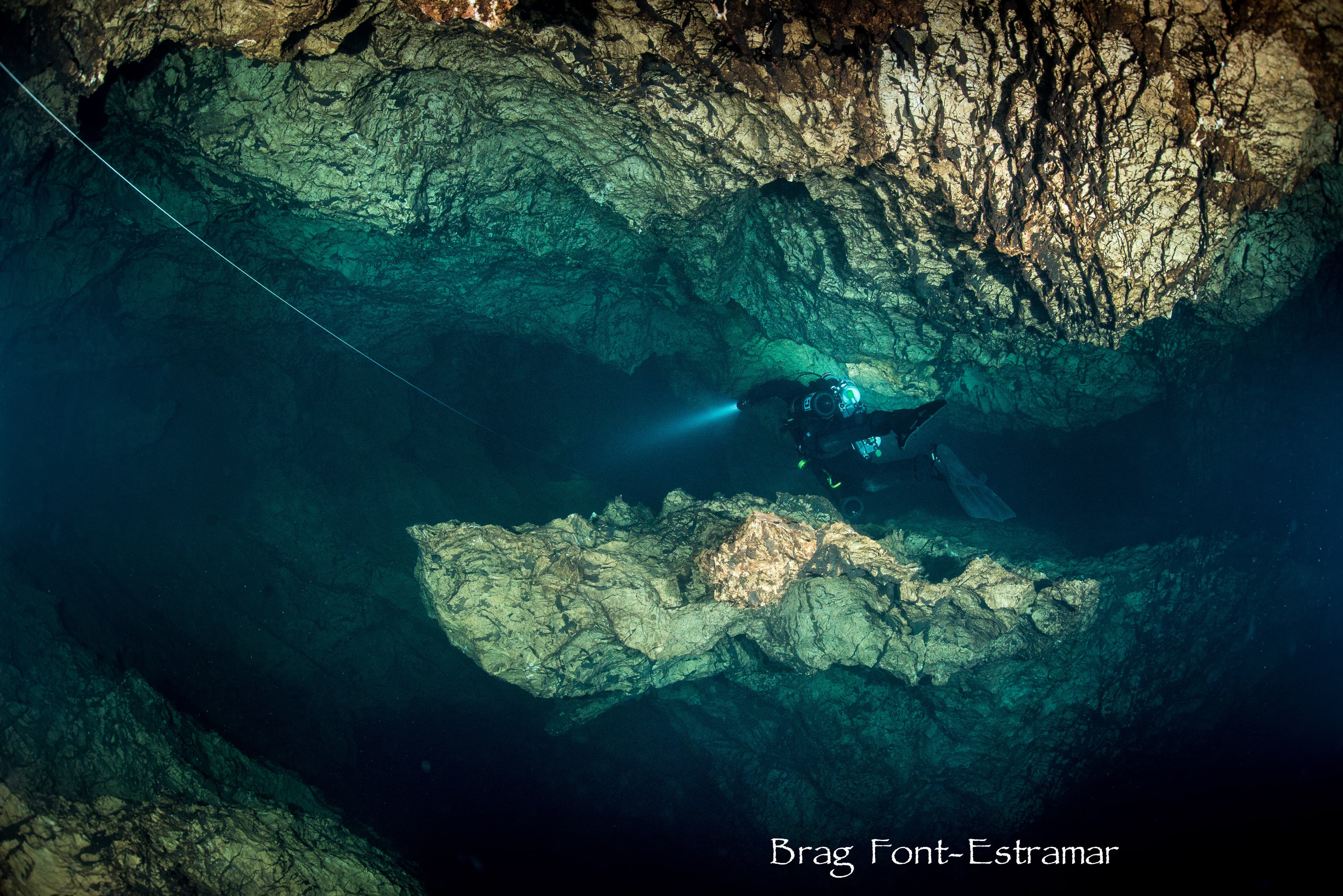 Cave diver following a guideline through a teal-lit underwater cave passage with rock formations