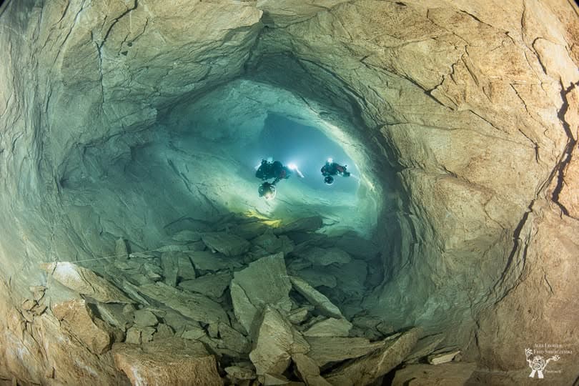 Two cave divers hovering in a large flooded chamber with boulders on the floor