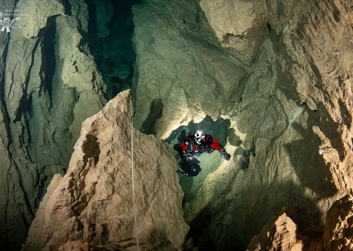 Lone cave diver dwarfed within a tall rocky shaft, a vertical guideline running down the rock face