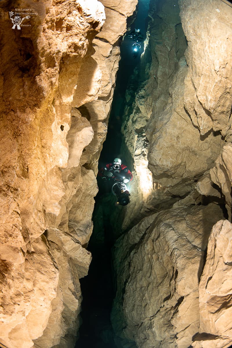 Two divers in a narrow vertical fissure between sheer rock walls, one above the other