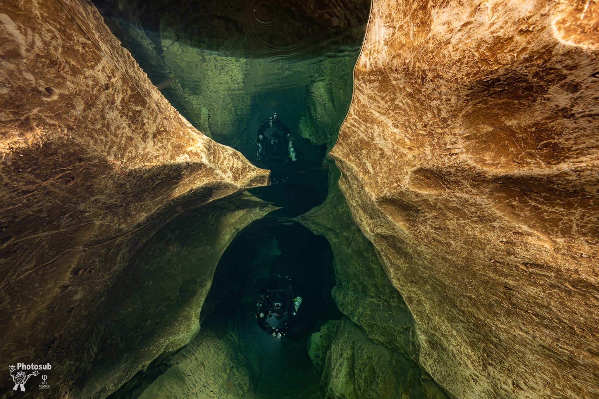 Two cave divers in a narrow passage reflected in a mirror-like water surface