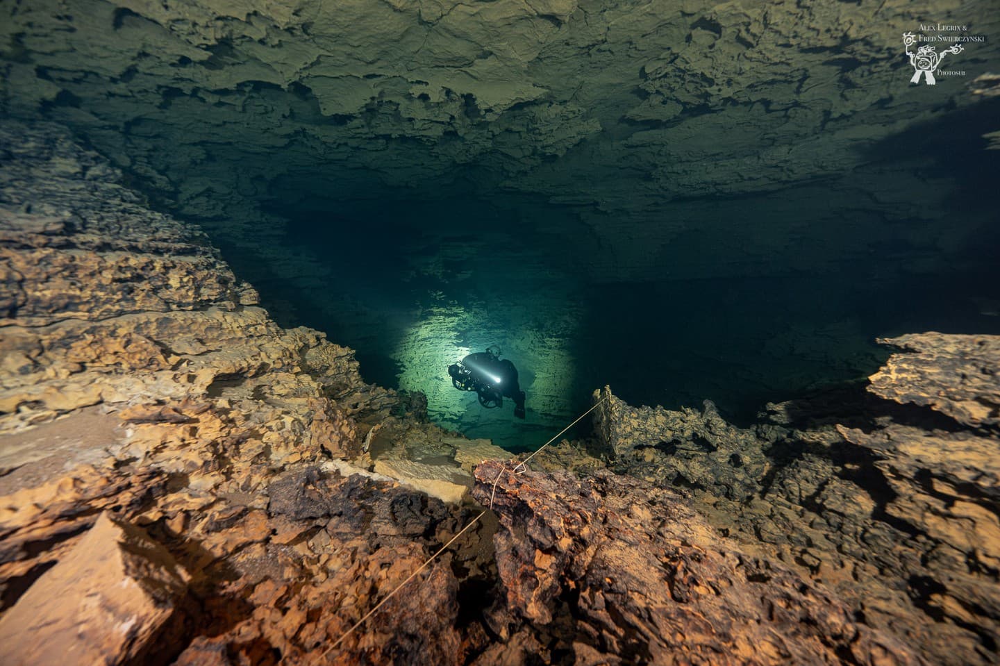 Wide view of a large cave chamber with rough rock formations, a distant diver lighting the passage and a guideline running across the rocks.