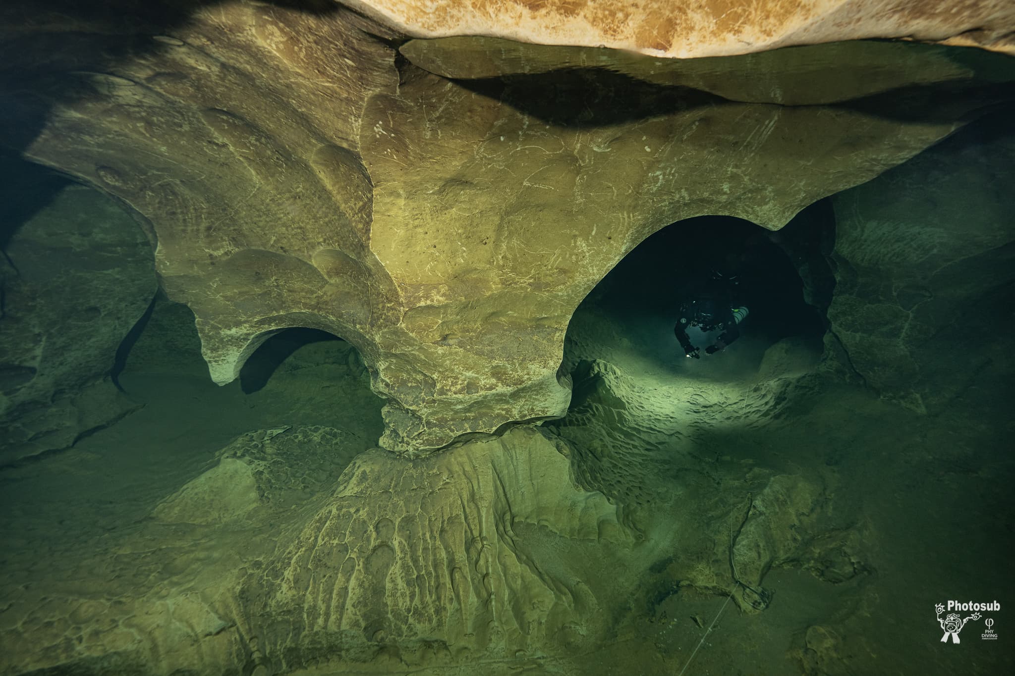 A wide cave passage with sculpted rock arches and a distant diver silhouetted in a side tunnel