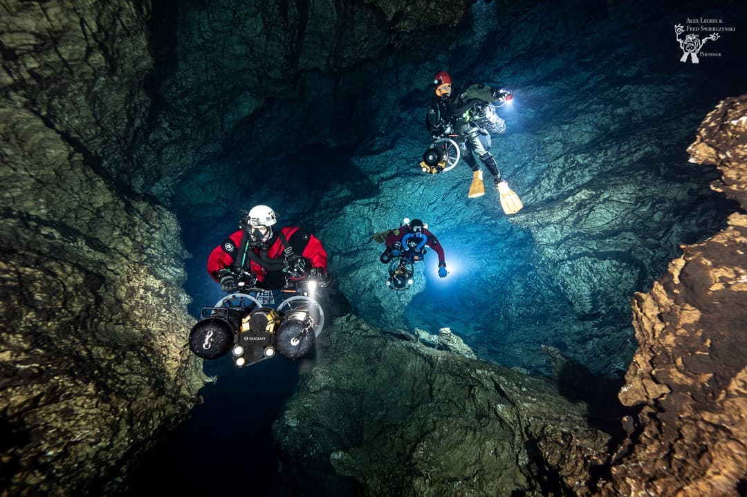 Three divers in dual sidemount rebreathers with DPVs in a rocky cave shaft