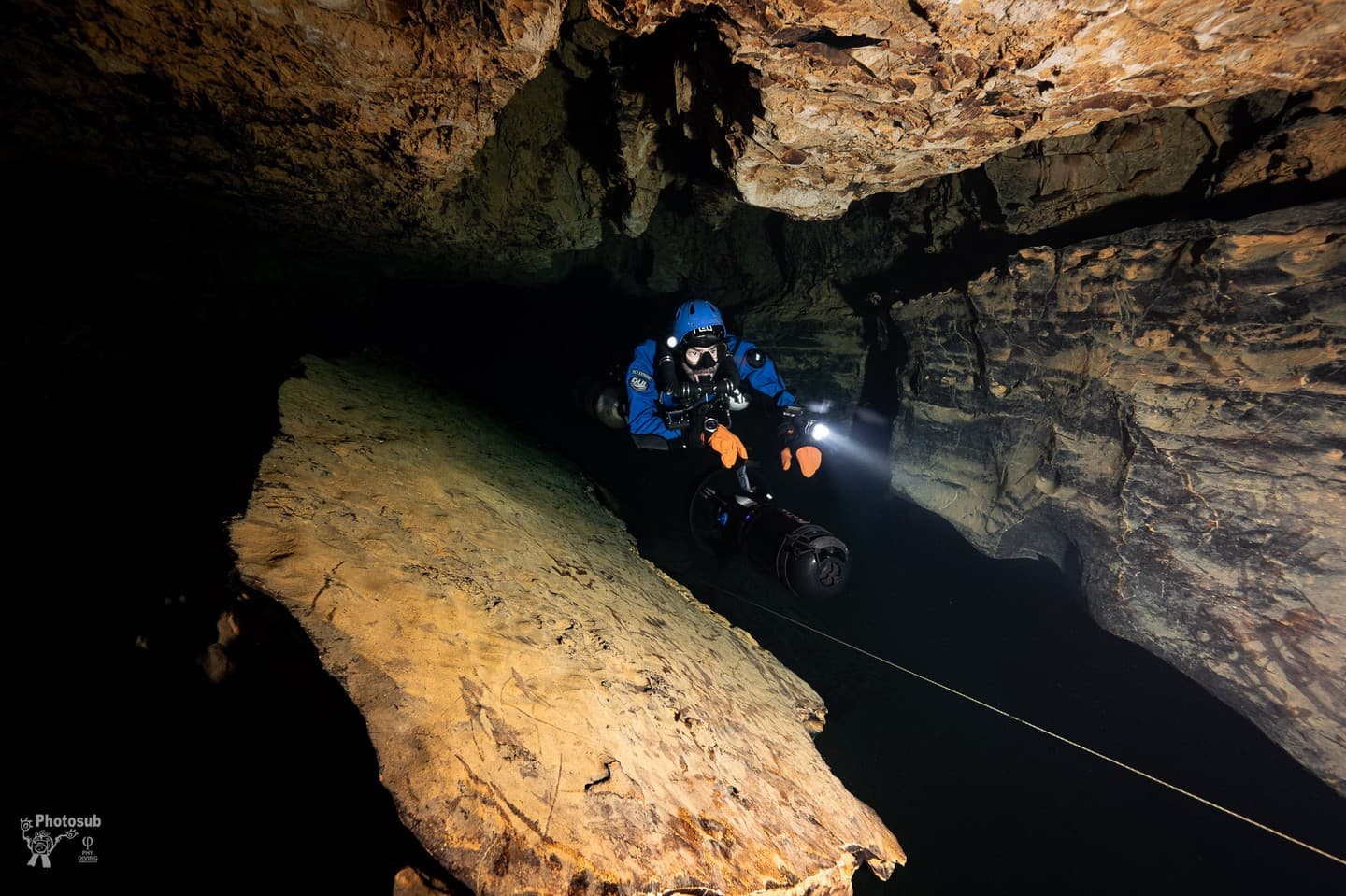 Diver in dual sidemount rebreathers along a cave wall with a DPV