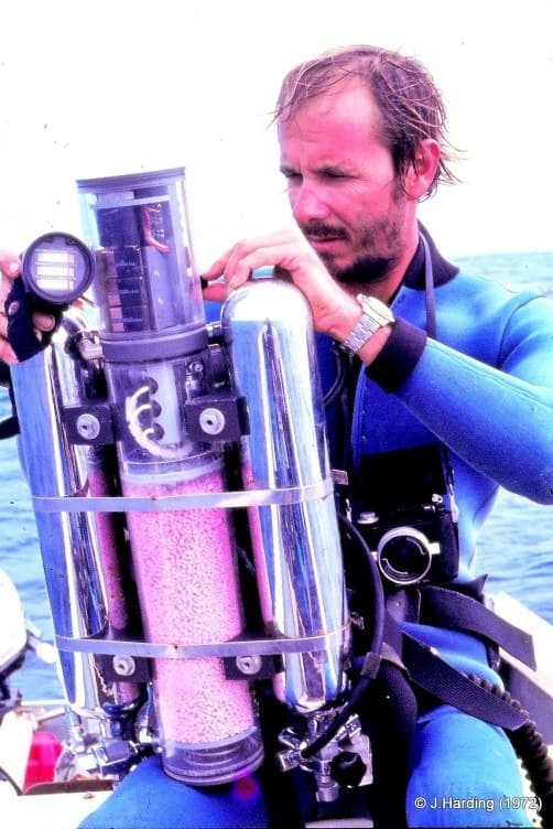 A diver in 1972 holding a chest-mounted rebreather with a transparent scrubber canister full of soda lime