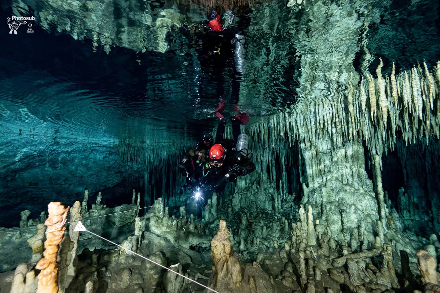 Two cave divers near the cavern ceiling following a guideline marked with a directional arrow among stalactites