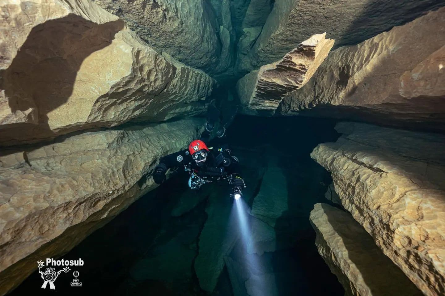 Rebreather diver with red helmet in a dramatic vertical cave shaft, light beam visible