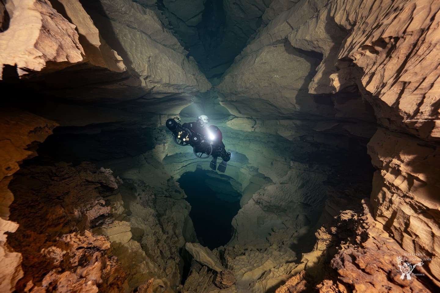 A rebreather diver hovering above a deep vertical pit in a large rocky cave chamber