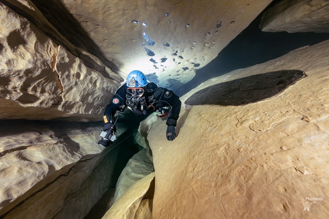 Close view of a rebreather diver in a blue helmet working through a narrow cave restriction over light-colored rock