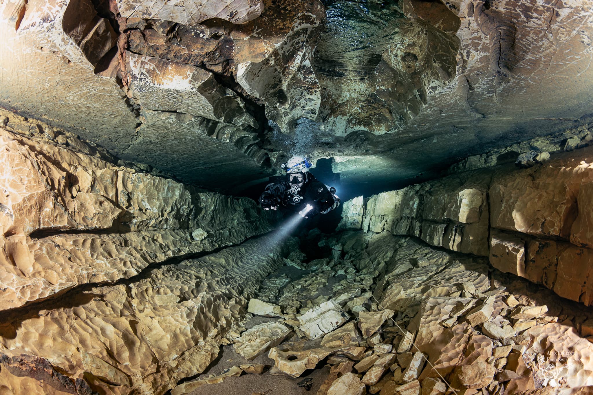 Open-circuit sidemount diver progressing along a wide low cave tunnel