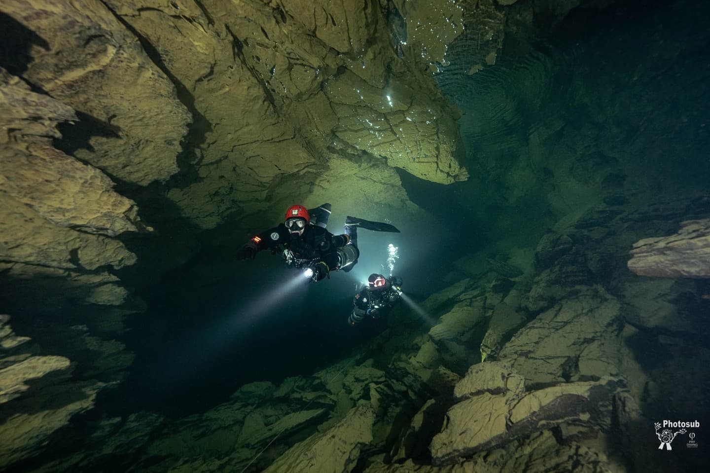 Two divers in horizontal trim swimming with primary lights through a dark rocky cave fissure