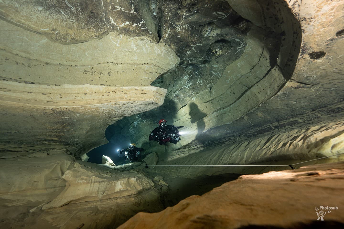 Two cave divers moving together along a guideline in a broad sculpted limestone gallery