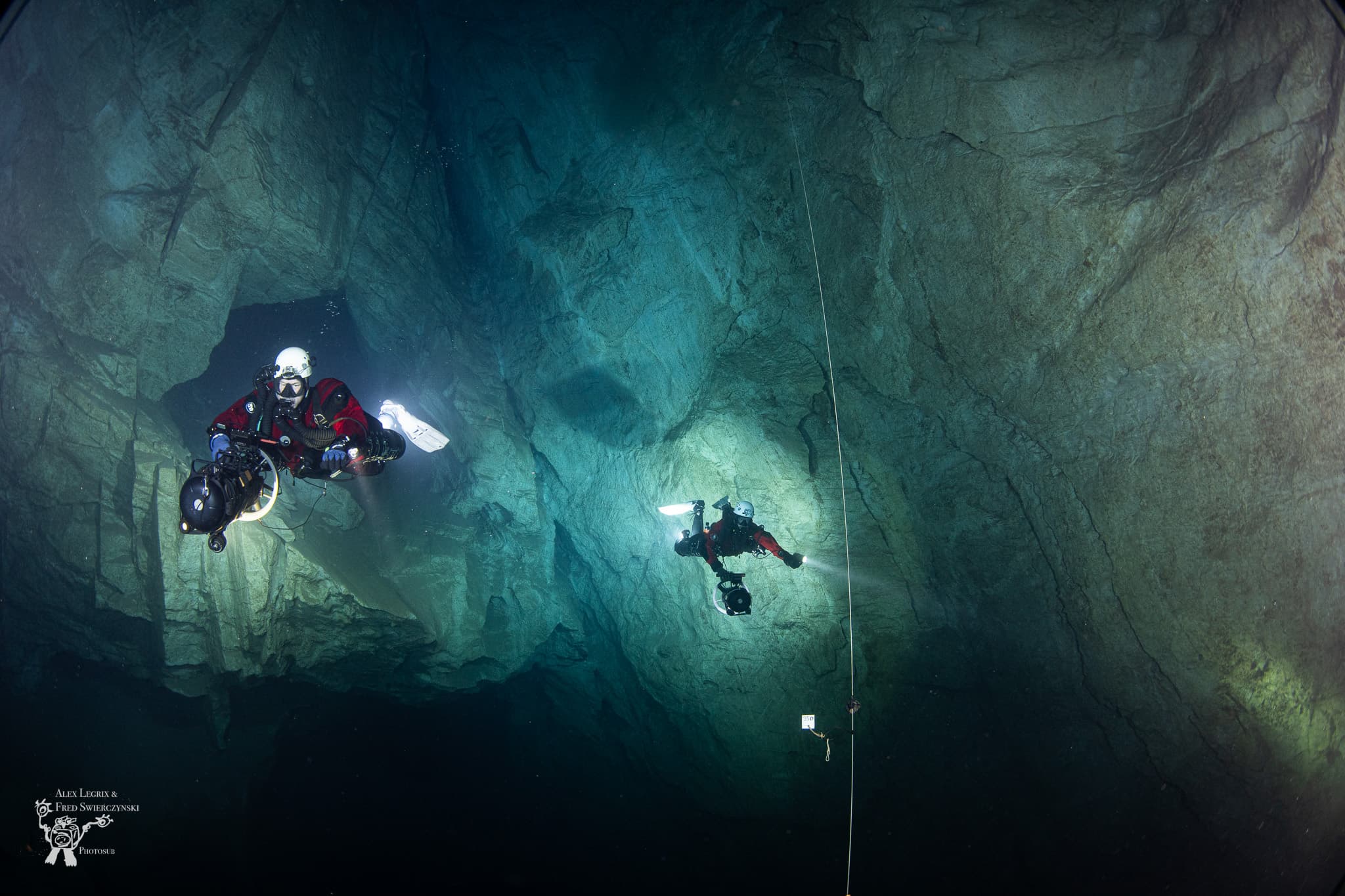 Two cave divers spaced apart in a vast dark chamber, a vertical guideline running between them with a spool on the floor